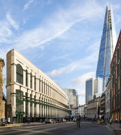 A city street with a curved modern building on the left and the tall glass Shard skyscraper in the background under a blue sky. People and cyclists are on the road.