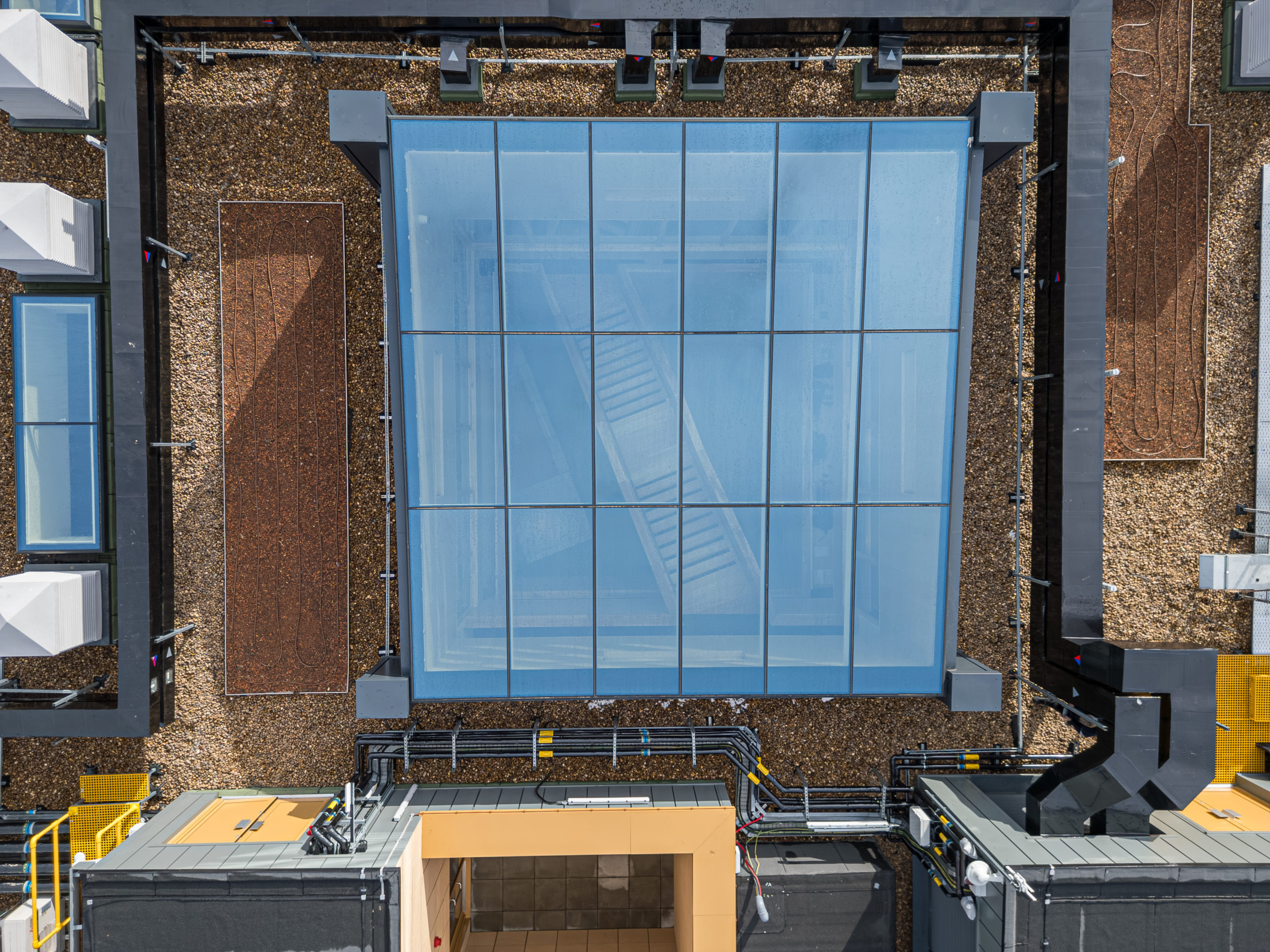 Aerial view of a building rooftop with a large multipart fixed rooflights rooflight at the centre, surrounded by gravel, vents, pipes, and rectangular structures.