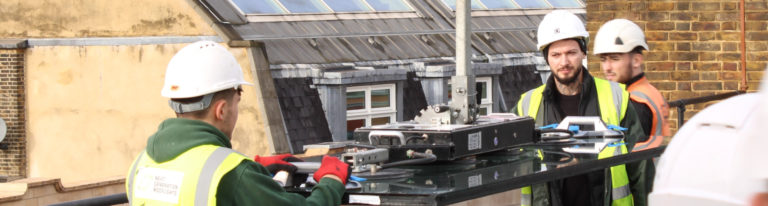 Three construction workers wearing safety vests and helmets are operating machinery on a rooftop, specifying sliding rooflights, with brick buildings in the background.